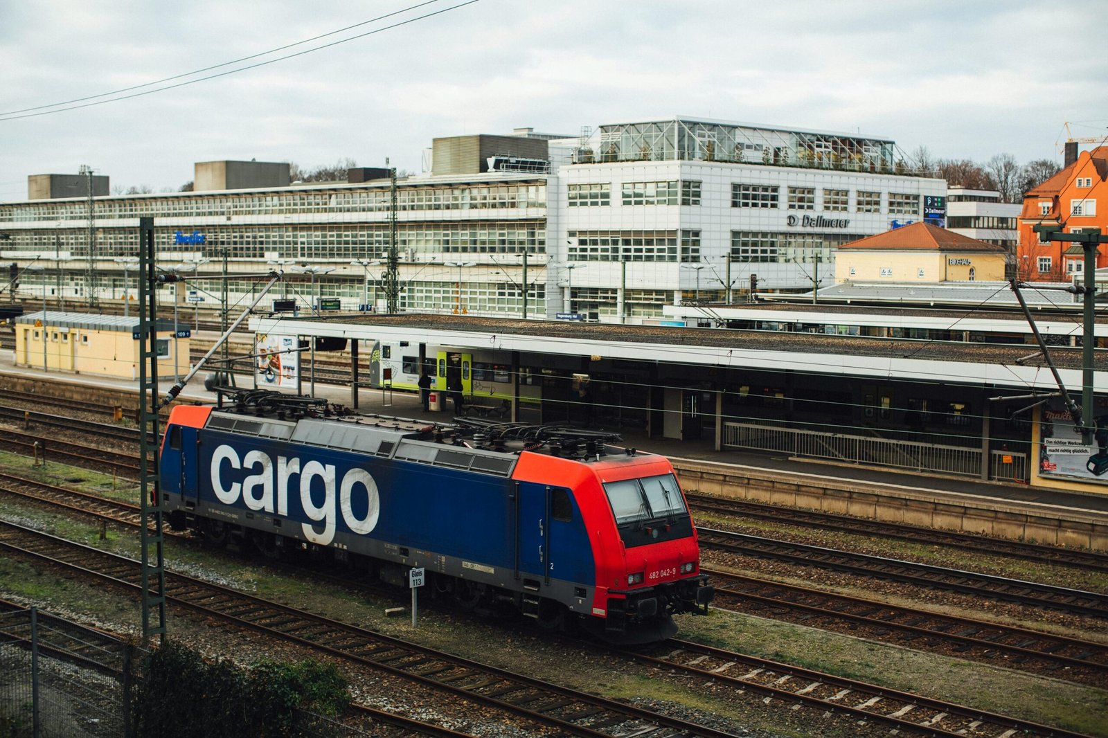 Cargo train parked at a modern urban railway station on a cloudy day.