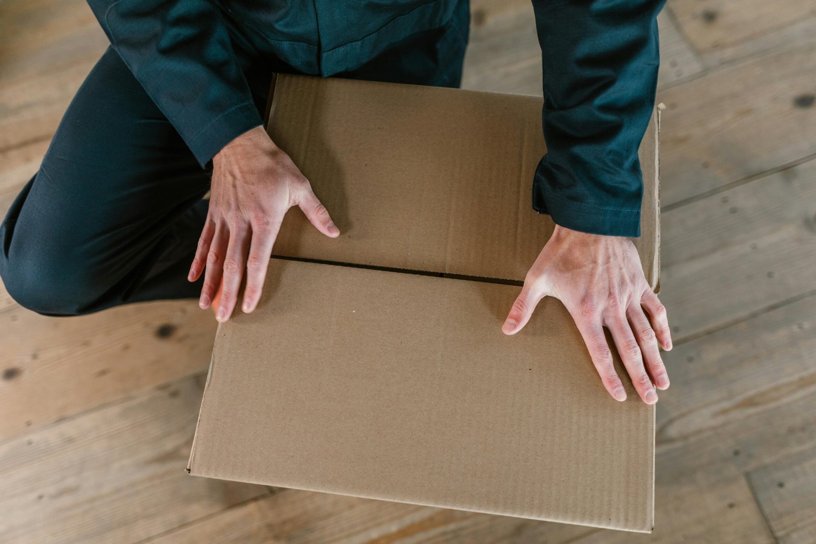 Close-up of hands holding a cardboard box on a wooden floor, captured from above.